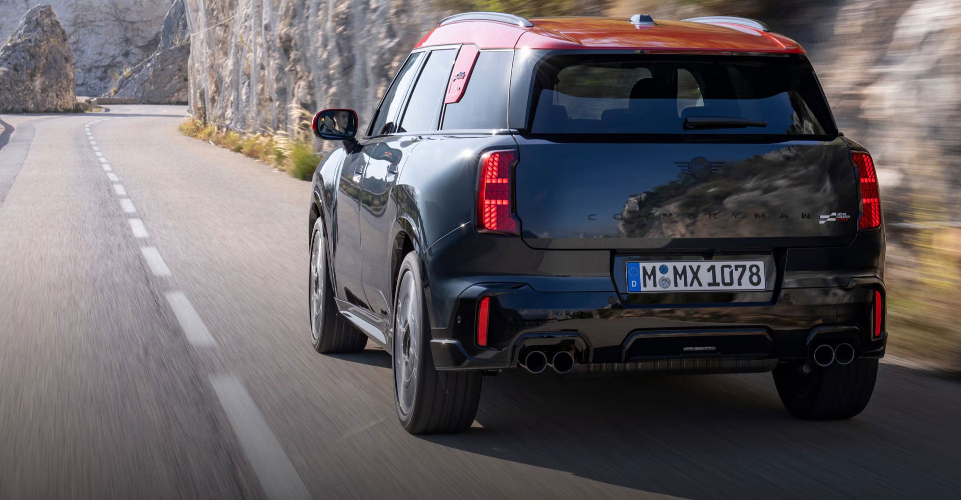Angled rear-to-front view of the MINI JCW Countryman ALL4 driving down a road surrounded by a wall of rock and foliage.